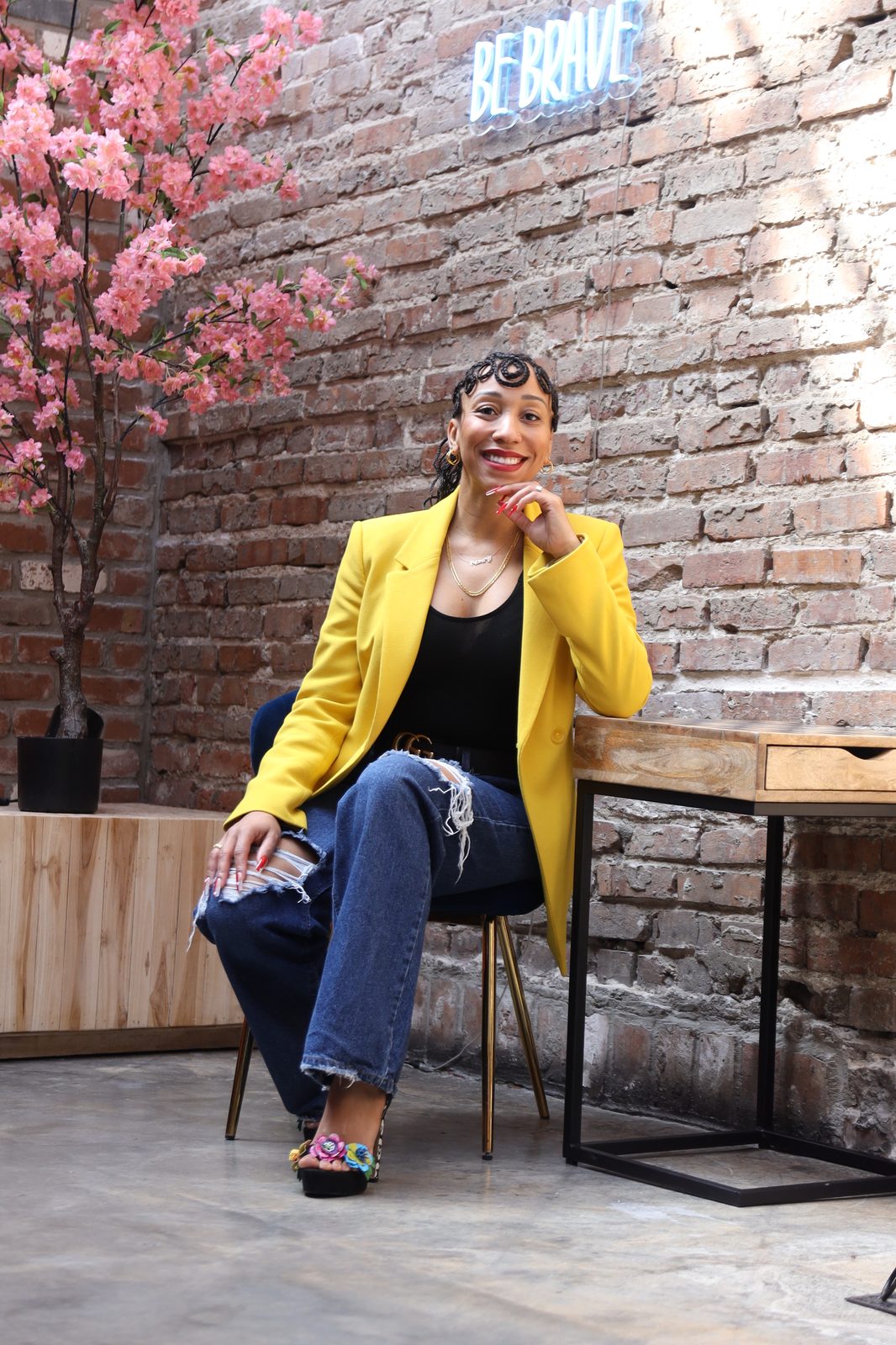 Kim Woodard sitting in a brick studio space under a 'Be Brave' neon sign.
